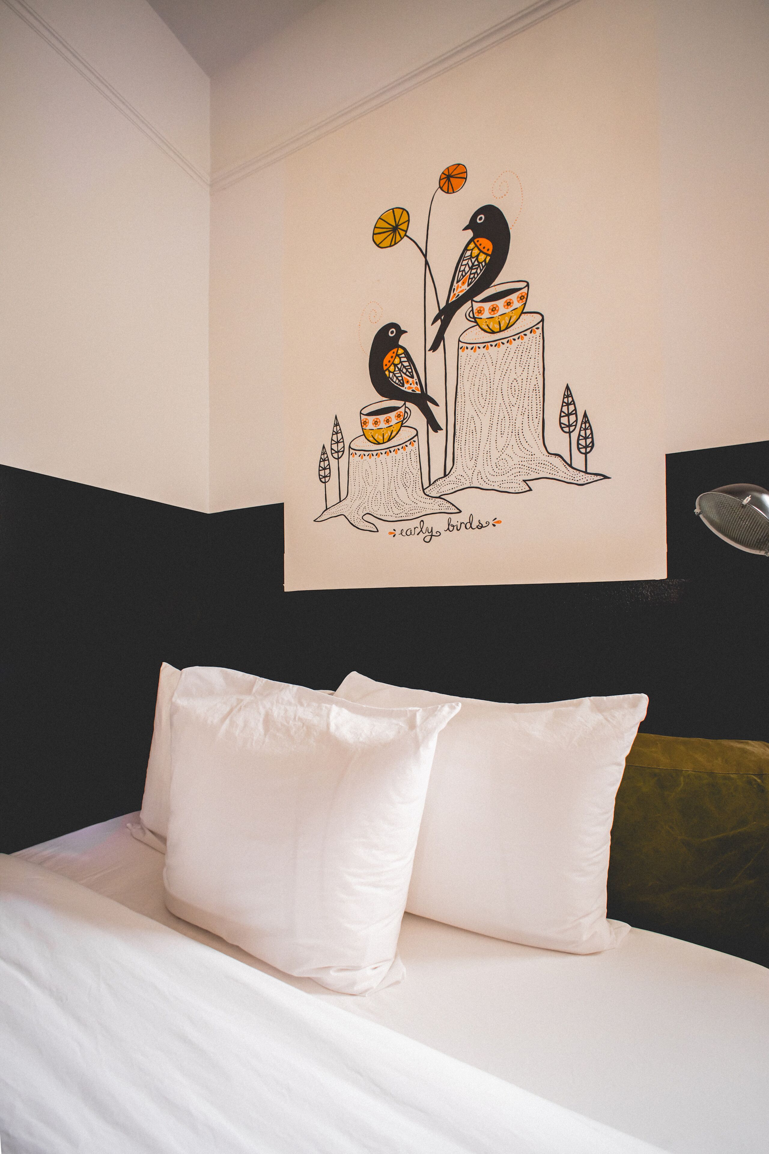 A bed with white bedding and two pillows, a wall with a drawing of birds and a cup, and a partial view of a headboard.