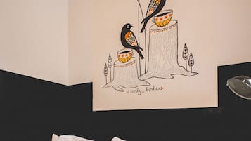 Una cama con ropa de cama blanca y dos almohadas, una pared con un dibujo de pájaros y una taza, y una vista parcial de un cabecero.