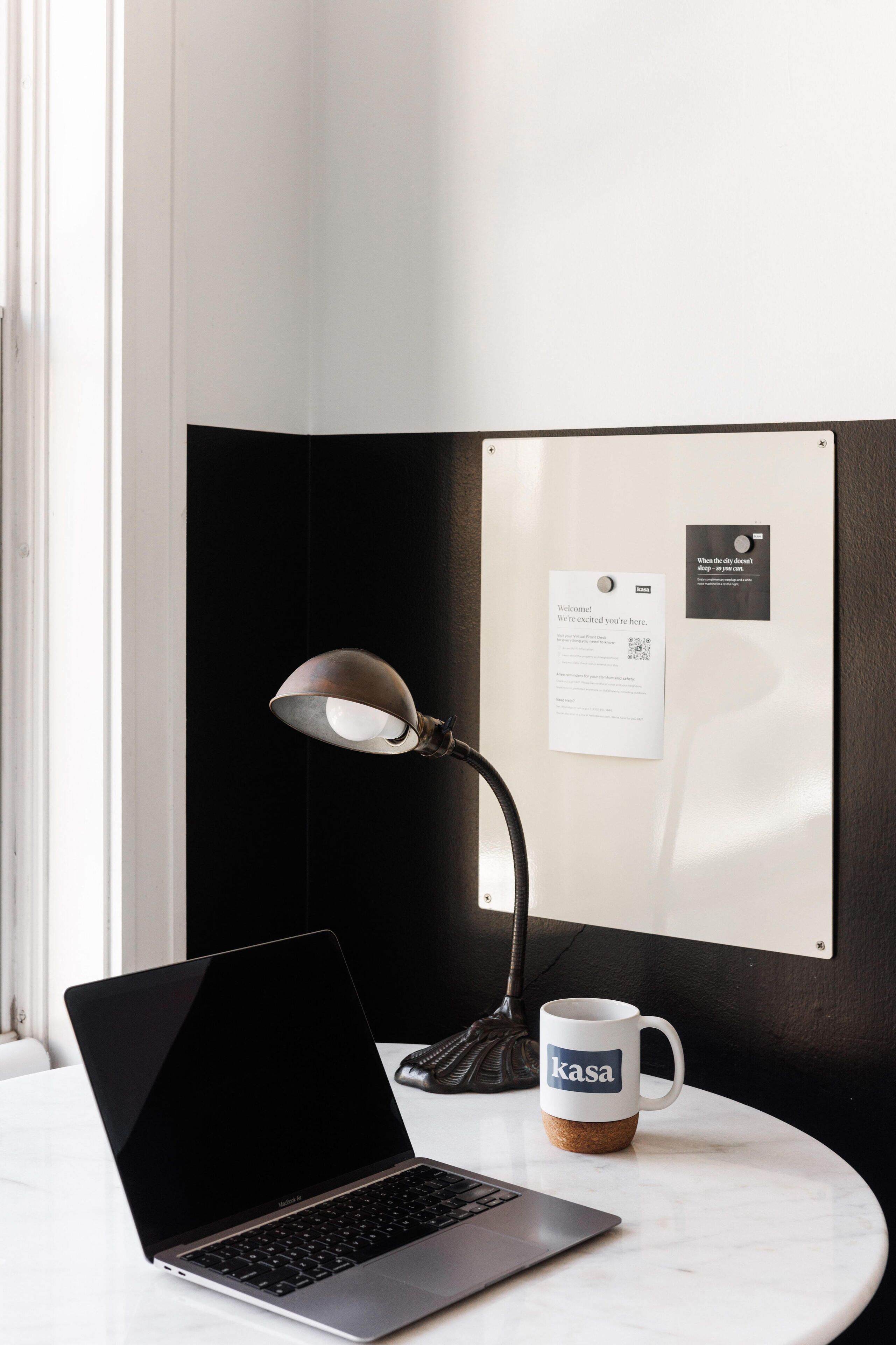A laptop on a marble table, a coffee mug with the word "kasa" on it, a desk lamp, and a bulletin board with a welcome sign and a business card.