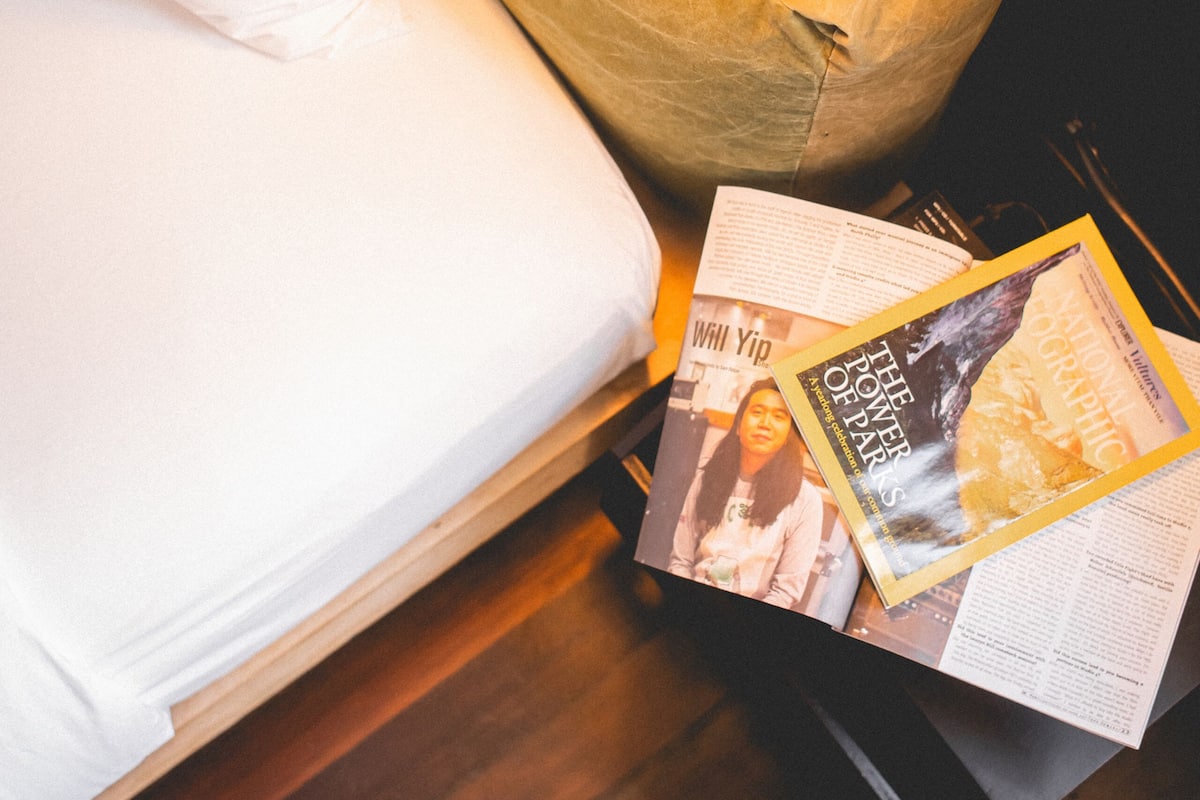 Een bed met een wit kussen en een geel hoofdeinde, een zwarte lamp, en tijdschriften op de houten vloer.