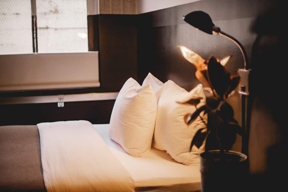 Een net opgemaakt bed met witte beddengoed en meerdere kussens, een bedlampje en een raam met jaloezieën.