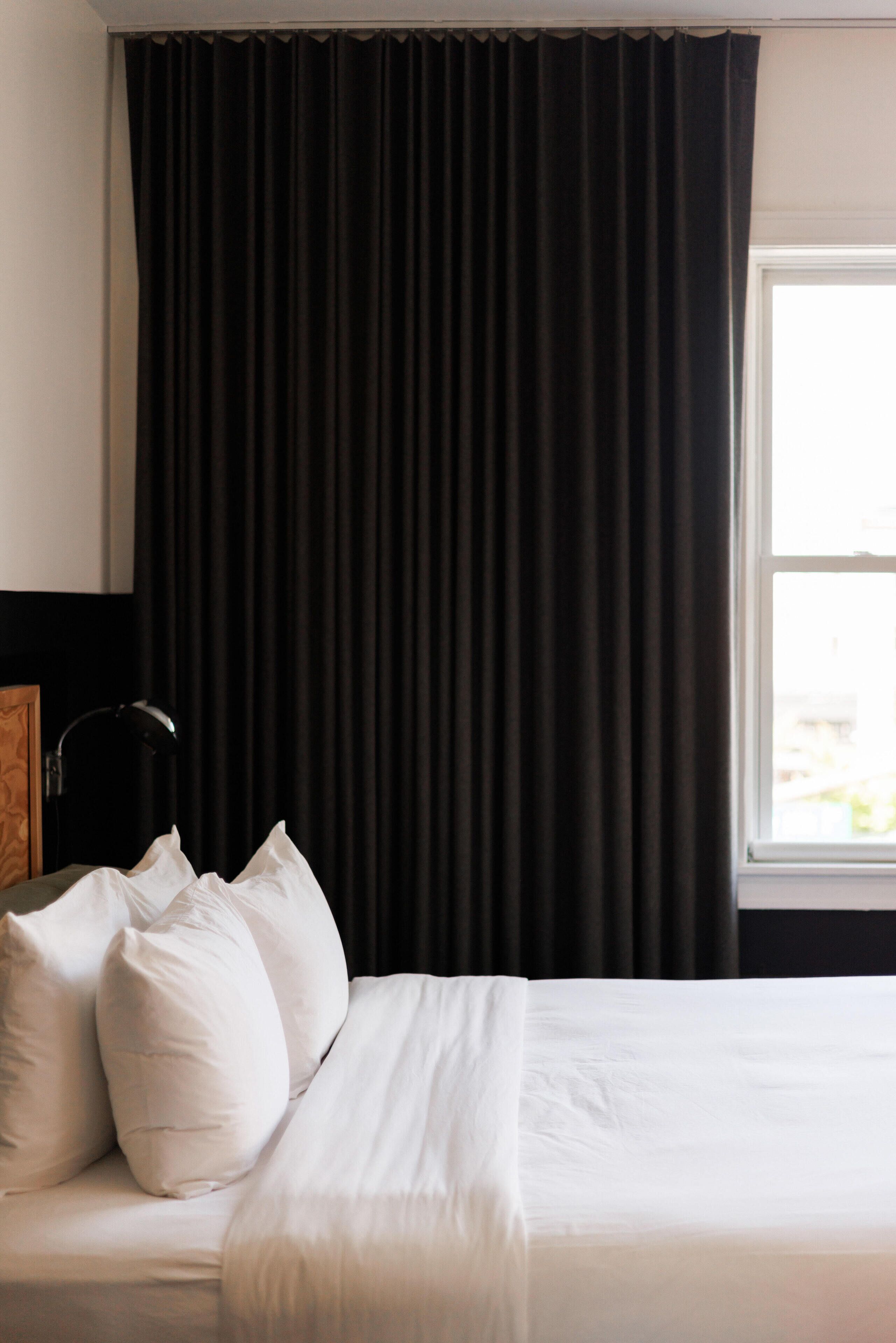 A hotel room with a bed, white pillows, a black lamp, and a window with curtains.
