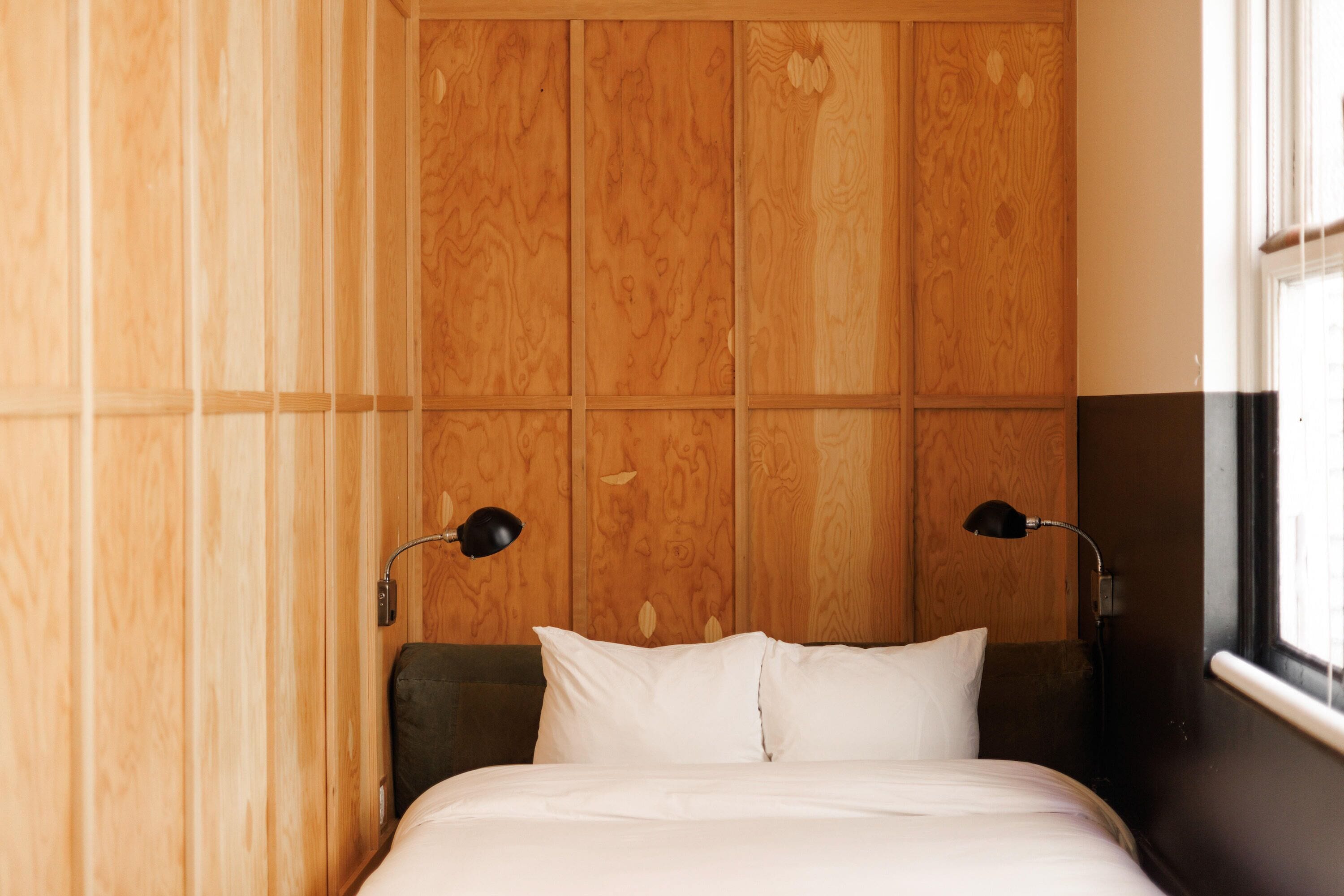 Una habitación con cabecera de madera, cama con ropa de cama blanca, dos lámparas de noche y una ventana a la derecha.