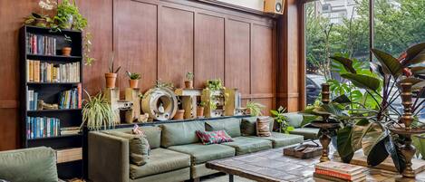 A cozy living room with a sectional sofa, a coffee table, a bookshelf, and various plants.
