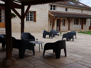 A covered outdoor seating area with modern chairs and tables, stone walls, and a traditional building with windows and a tiled roof. - Sleeps 13/15, 270 m2 house with swimming pool / CHEQUES ANCV ACCEPTES (Bonnes)