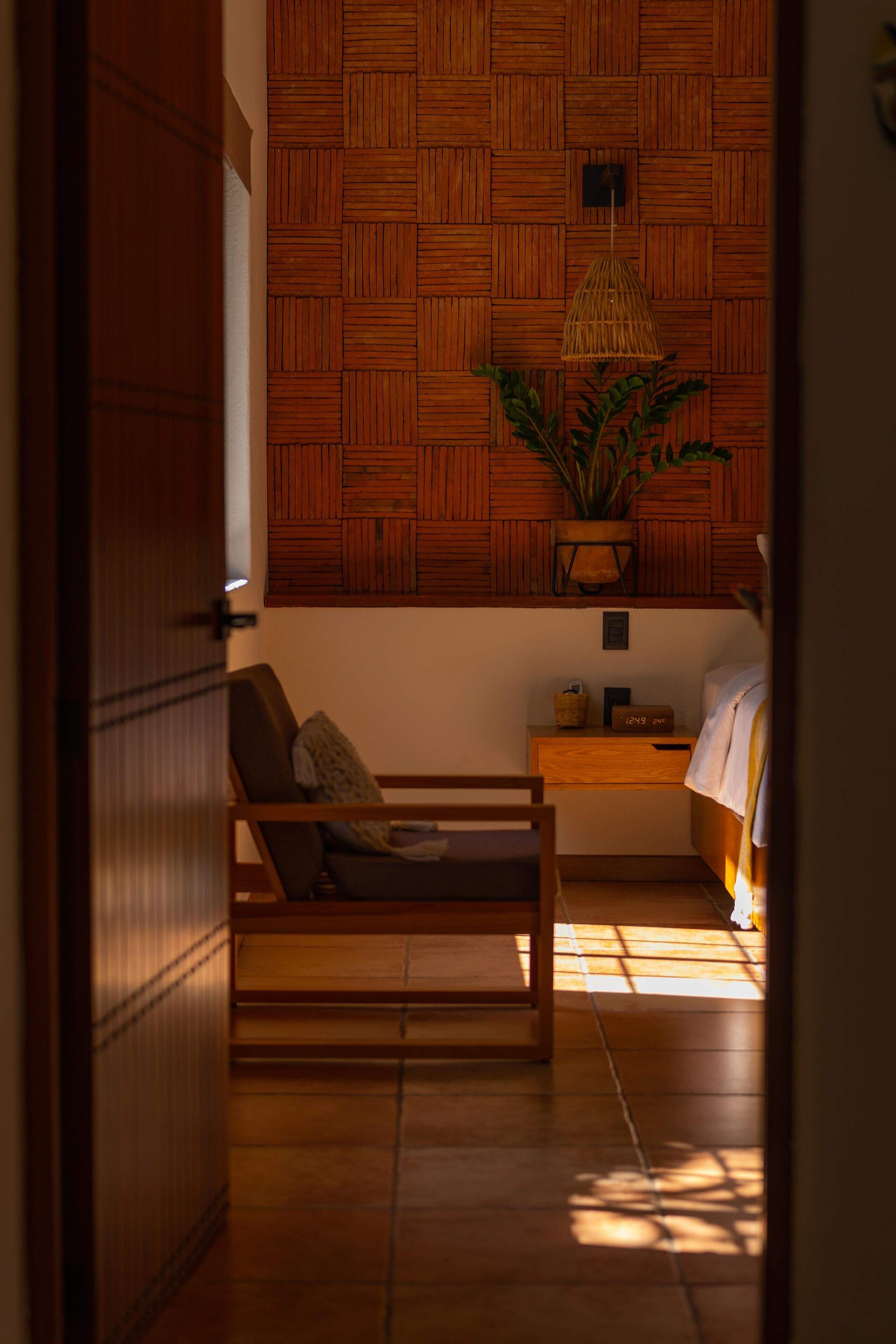 Una habitación con un panel de madera, una lámpara de mimbre, una planta, un banco de madera, una mesita de noche y una cama.