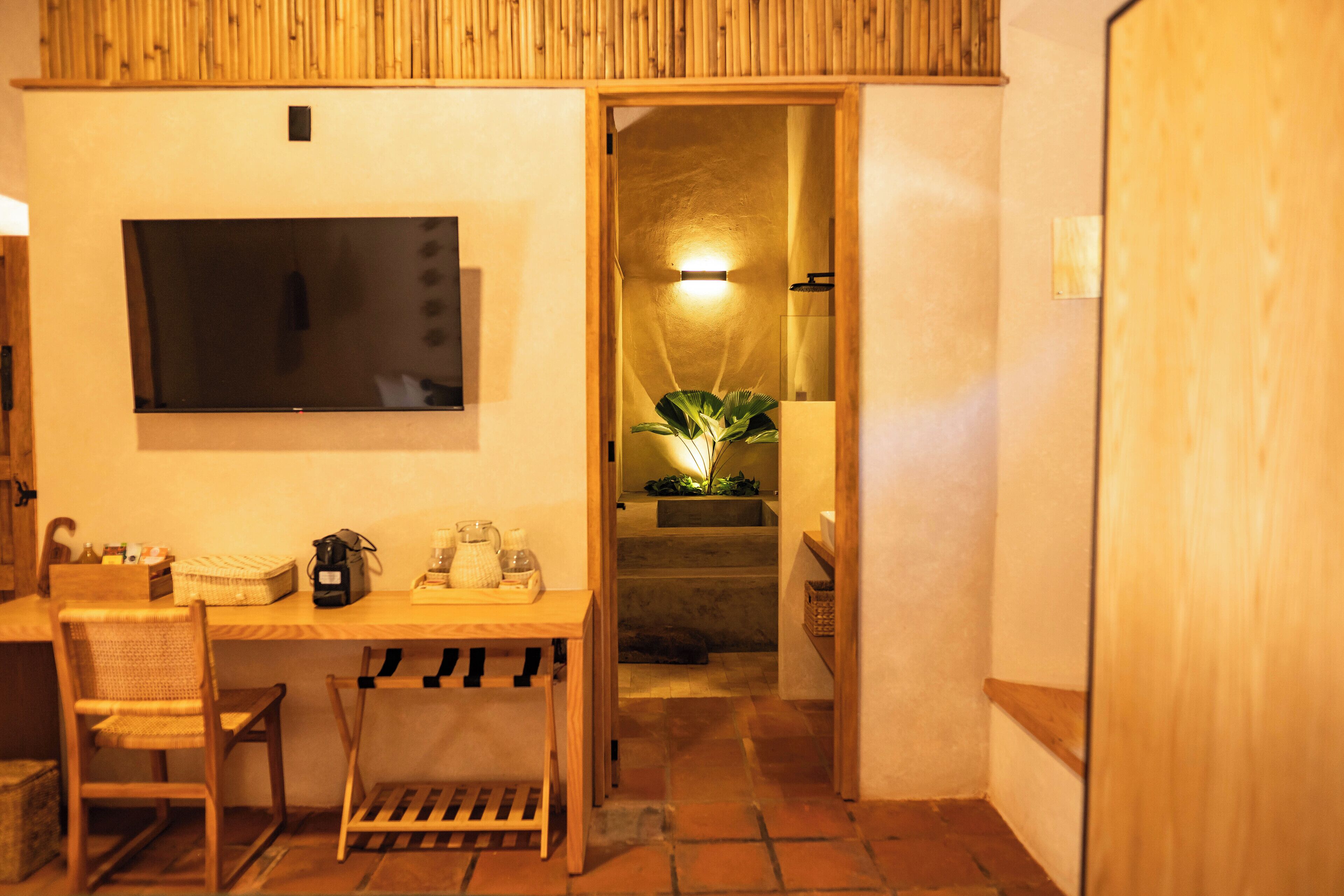 Habitación con escritorio de madera, una silla y un televisor montado en la pared. Se ve una escalera que lleva a un baño con una planta y una lámpara.