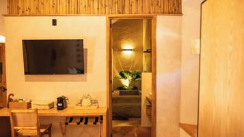 Habitación con escritorio de madera, una silla y un televisor montado en la pared. Se ve una escalera que lleva a un baño con una planta y una lámpara.