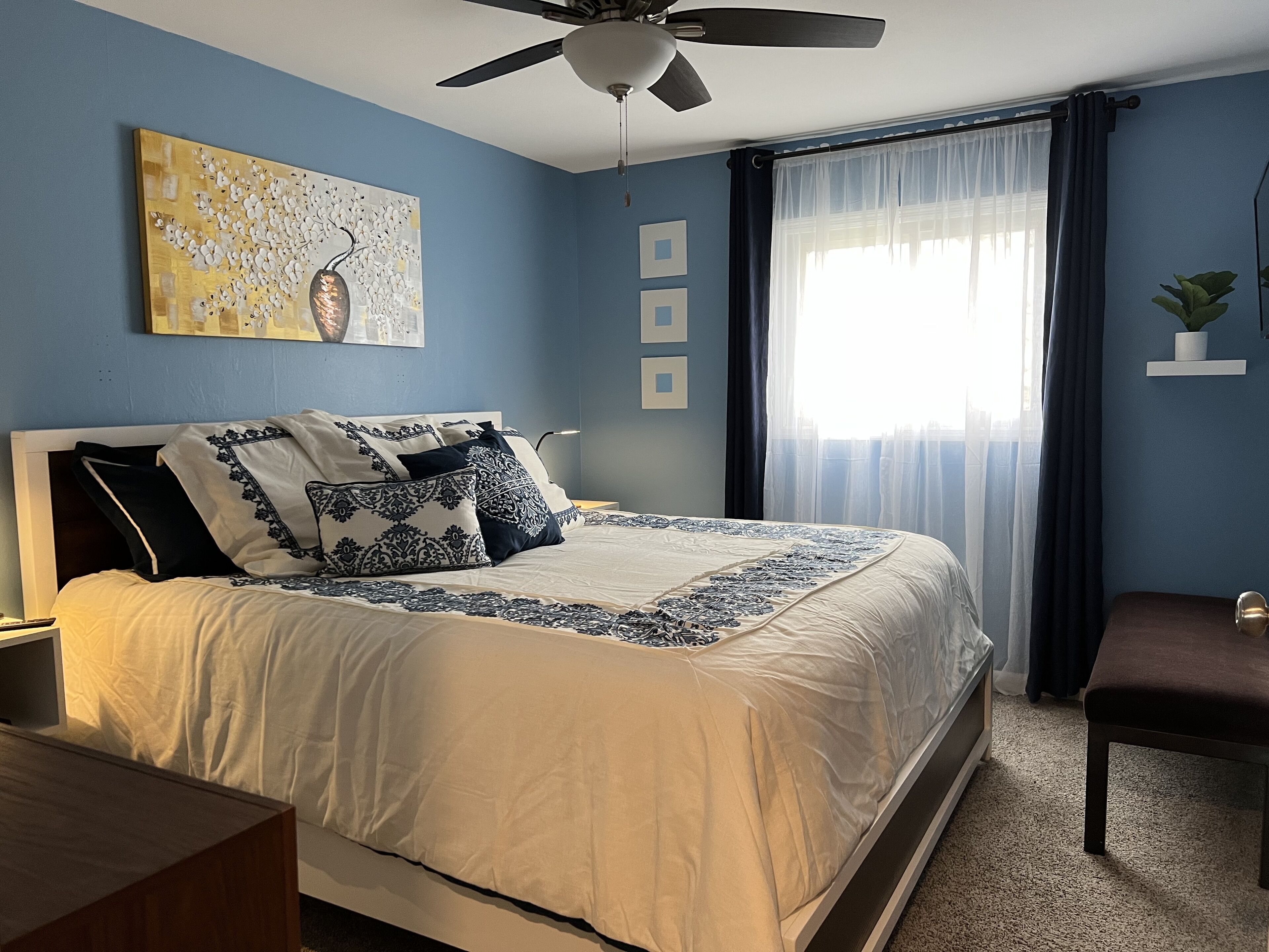 A bedroom with a bed, a ceiling fan, a window with curtains, a wall art piece, and a small potted plant.