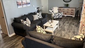 Un salón con un sofá gris oscuro, una mesa de centro blanca y cojines estampados. Hay un aparador blanco con cajones y un espejo en la pared.