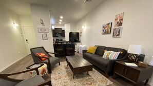 Een moderne woonkamer met een bank, fauteuils, een salontafel en een vloerkleed. In de achtergrond is een keuken te zien.