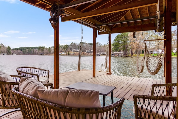 Una terraza de madera con muebles de mimbre y una hamaca, con vista a un lago y casas al fondo.