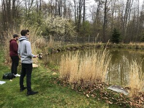 Zwei Personen angeln an einem Teich, umgeben von Bäumen und hohem Gras.