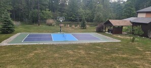 Un terrain de tennis extérieur doté d’un panier de basketball, entouré d’une pelouse et d’arbres.