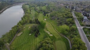 En vy över en golfbana från ovan, med en flod, stadens silhuett och ett bostadsområde i bakgrunden.