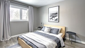 Ett modernt sovrum med en säng, ett sängbord, en liten avlastningsyta, ett fönster med gardiner och en inramad tavla på väggen.