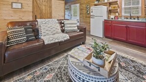 Un salon chaleureux avec un canapé en cuir marron, des coussins à motifs et un tapis tissé. Une table basse en bois porte une plante et des livres. La cuisine est dotée de meubles rouges et d’un réfrigérateur blanc.