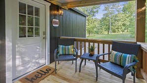 Un porche avec deux fauteuils en osier et une petite table, un paillasson avec l’inscription « Bonjour » et une pancarte « Bienvenue ».