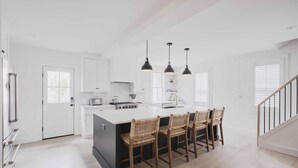 Una cocina moderna con armarios blancos, una isla central con taburetes de bar, iluminación colgante y una escalera a la derecha.