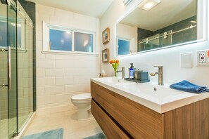 Un baño moderno con cabina de ducha de vidrio, inodoro blanco, tocador de madera con lavamanos blanco y un espejo grande.