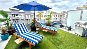 Una terraza en la azotea con dos sillones de descanso a rayas, una parrilla y plantas en macetas.