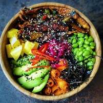 Un tazón de madera lleno de diversos alimentos, como carne, aguacate, edamame y verduras.