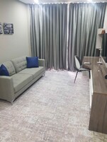 Una sala de estar con un sofá gris, cojines azules, una mesa de centro de madera, una silla, un televisor y una cortina.