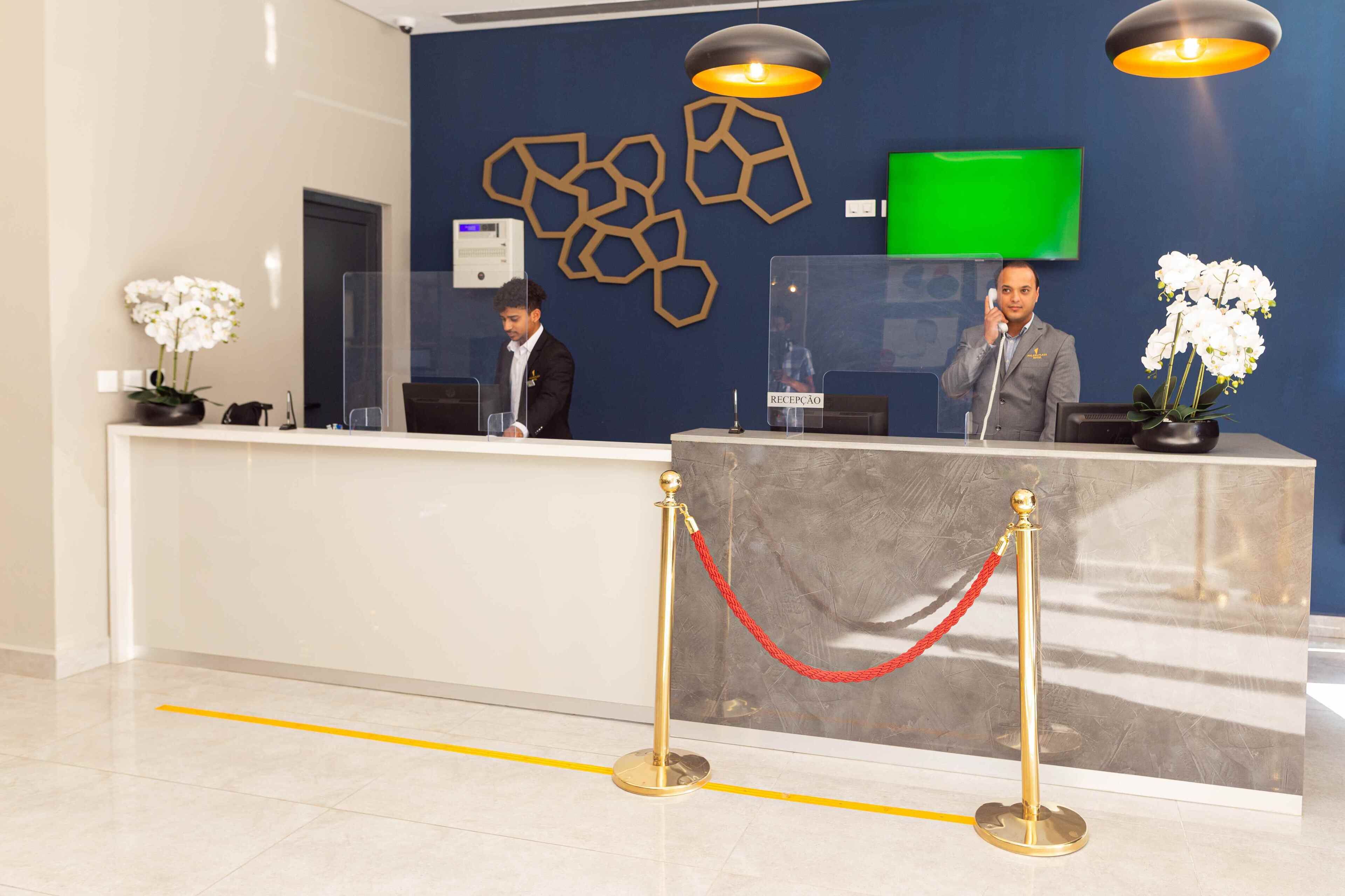 Un mostrador de recepción con dos recepcionistas, una pantalla verde y una barrera de cristal transparente.