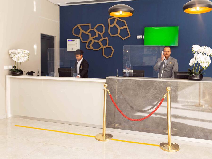 Un mostrador de recepción con dos recepcionistas, una pantalla verde y una barrera de cristal transparente.
