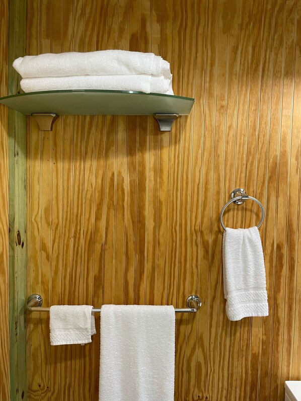Une salle de bain avec un lambris en bois, une étagère en verre o ù sont pliées des serviettes, et un porte-serviettes en métal sur lequel est suspendue une serviette.