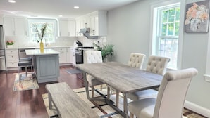 A modern kitchen with white cabinets, a central island, and a dining area with a wooden table and white upholstered chairs.