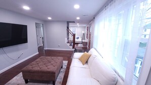 A living room with a flat-screen TV mounted on the wall, a brown ottoman, a white sofa with a yellow pillow, and a large window with sheer curtains.