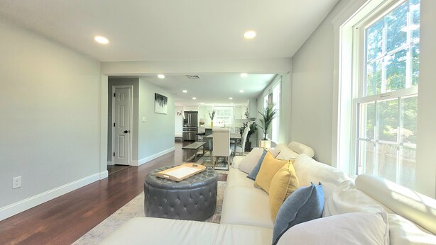 A spacious living room with a large window, a dark ottoman, and a white sofa with cushions.
