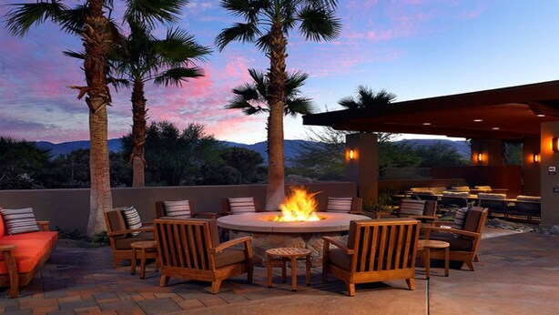 Un espacio al aire libre con zona de fogata, sillas de madera y vista a montañas y palmeras al atardecer.
