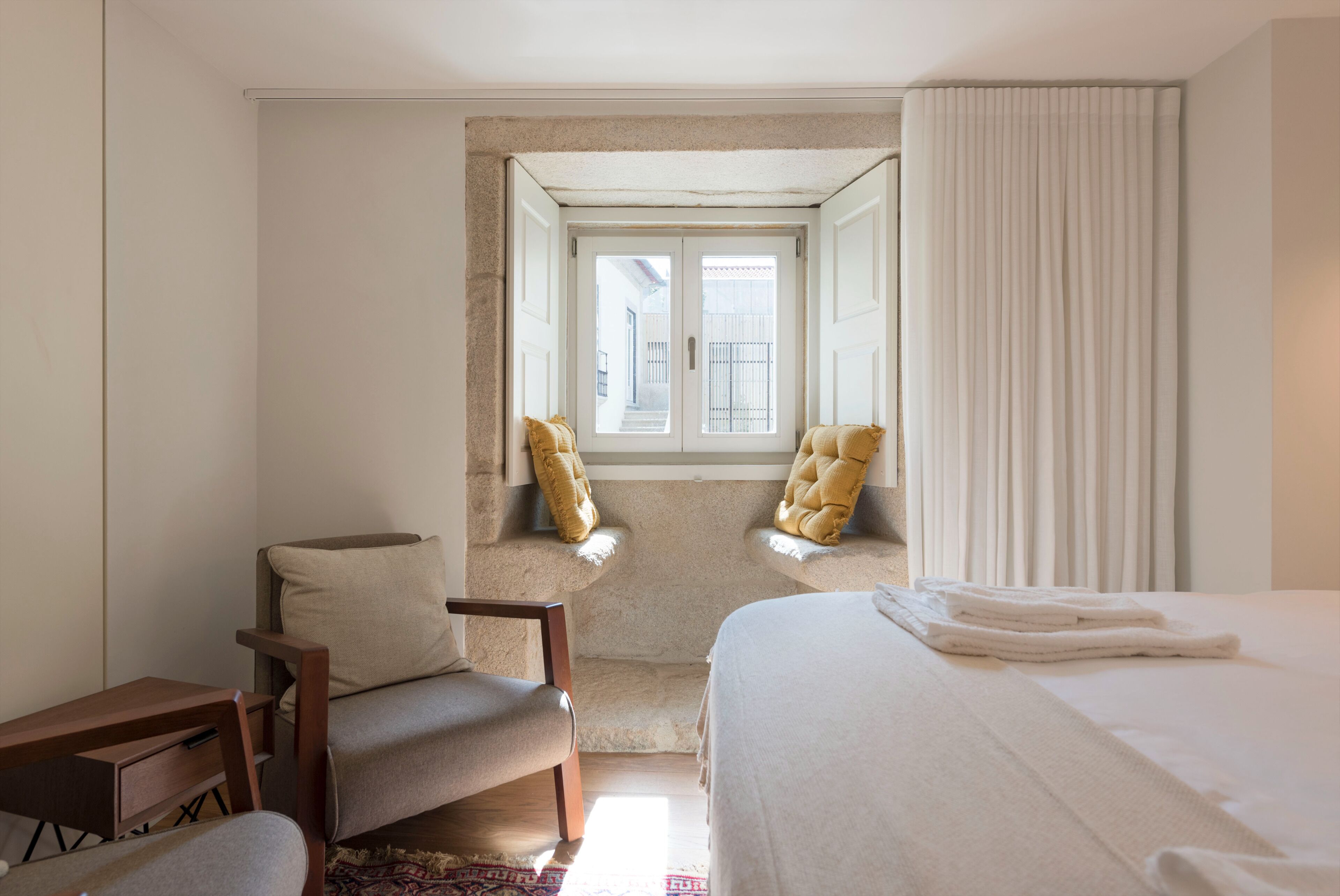 Une chambre d’hôtel avec un lit, deux fauteuils, un petit bureau et une fenêtre avec des rideaux.