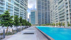 Un espace moderne aménagé au bord de la piscine, avec des chaises longues et une piscine à l’eau d’un bleu limpide, le tout entouré d’immeubles de grande hauteur.