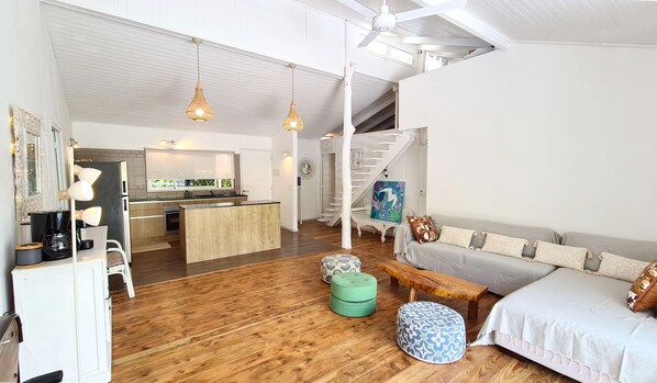 Un vaste espace de vie doté d’un plancher en bois, d’un canapé, de poufs, d’un banc et d’une cuisine en arrière-plan.