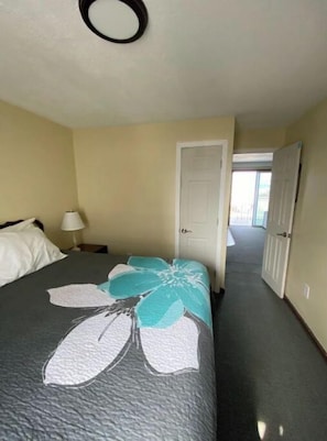 Quarto de hotel com uma cama, mesa de cabeceira, luminária e porta que dá para outro cômodo.