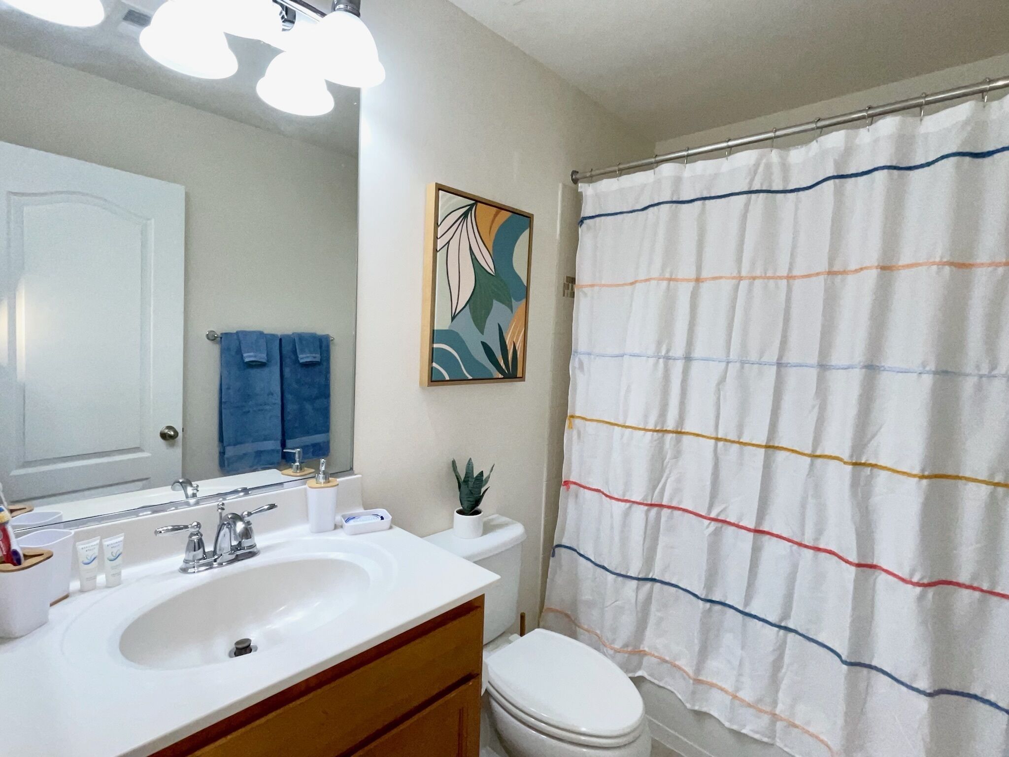 Een badkamer met een witte wastafel, spiegel en toilet. Aan de muur hangt een blauwe handdoek en er is een kleurrijk douchegordijn.