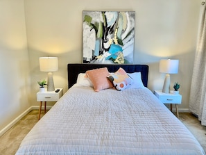 A neatly made bed with a quilted cover, two bedside tables with lamps and small plants, a large abstract painting on the wall, and a window with curtains.