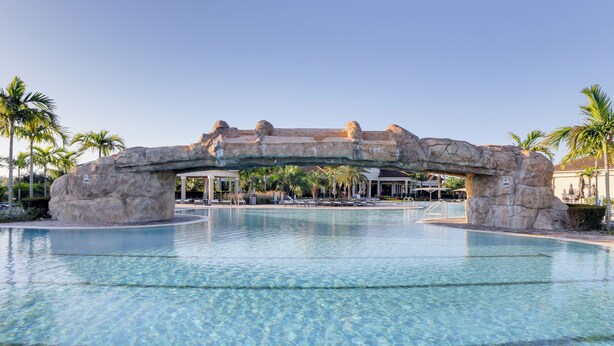 A clear pool with a rock archway, palm trees, and lounge chairs.