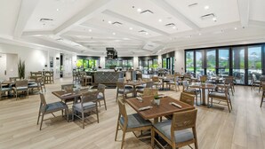 A spacious dining area with multiple tables and chairs, a central bar area, and large windows offering views of the outdoors.
