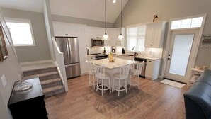 A modern kitchen with white cabinets, a stainless steel refrigerator, a microwave, and a dining area with white chairs. - Steps from the beach and Pool #62 (Osoyoos)