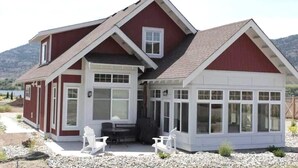 A two-story house with a large bay window, a covered patio with chairs, and a view of a lake. - Steps from the beach and Pool #62 (Osoyoos)