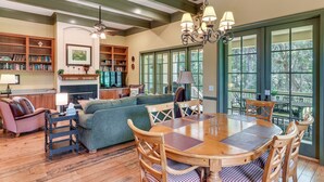A spacious living room with a fireplace, a dining area with a round table and chairs, and a large window with a view of trees. - 5 Mi to Old Town Bluffton: Cottage w/ Patio (Bluffton)