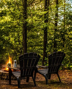 A forest scene with two Adirondack chairs and a metal table with a kettle and mug.