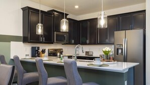 A modern kitchen with dark cabinets, a large island with bar stools, stainless steel appliances, and pendant lighting. - Charming Villa | Themed Rooms | Prime Location (Four Corners)