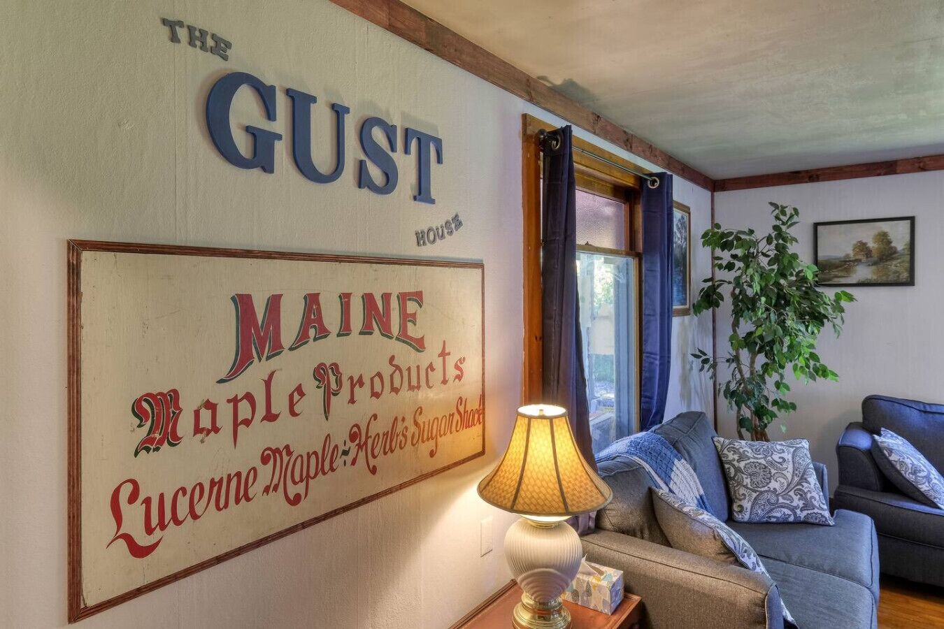 Ett mysigt vardagsrum med en stor skylt som visar texten "THE GUST" och "MAINE Maple Products". En lampa och en soffa syns i rummet.