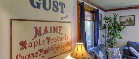 Ett mysigt vardagsrum med en stor skylt som visar texten "THE GUST" och "MAINE Maple Products". En lampa och en soffa syns i rummet.