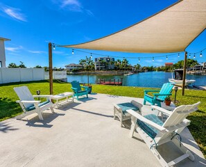Un patio al aire libre con sillas Adirondack blancas y turquesas, una mesa de hormigón y un toldo que proporciona sombra.
